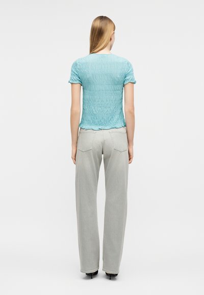 Γυναίκα με μακριά ξανθιά μαλλιά, φορώντας ένα υφασμάτινο ανοιχτό μπλε μπλουζάκι με κοντά μανίκια και ανοιχτό γκρι τζιν παντελόνι με ίσια γραμμή, όρθια με την πλάτη στραμμένη.