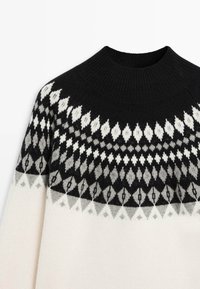 Gebreide trui in crèmekleur, met een zwart, grijs en wit geometrisch patroon bij het schouderstuk. Hoge zwarte geribde halslijn. Zachte textuur.
