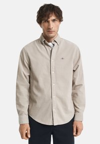 Man in een beige corduroy overhemd met knopen over een wit onderhemd, gecombineerd met zwarte broek, staand tegen een effen achtergrond.