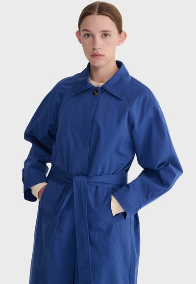 Jeune femme portant un trench-coat bleu ceinturé avec col, debout mains dans les poches devant un fond clair uni.