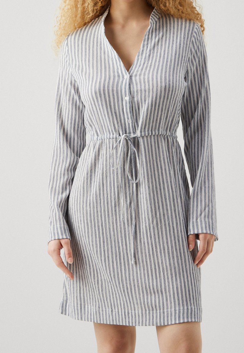 Femme portant une robe boutonnée jusqu'aux genoux, manches longues, avec des rayures verticales bleues et blanches et une ceinture nouée à la taille.