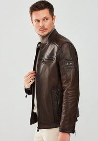 Brown leather jacket featuring zippered pockets, ribbed texture at shoulders, and a fitted design with a standing collar.