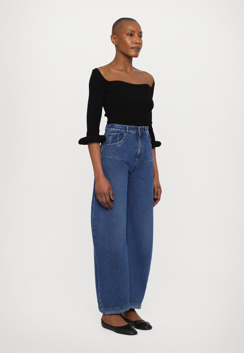 Top nero a coste scollato sulla spalla con maniche lunghe; jeans blu a vita alta e a gamba larga; scarpe nere basse; design minimalista e tessuto texture.