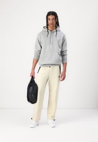 Sweatshirt cinzento com capuz e bolso frontal, calças de treino creme afuniladas, mochila preta e ténis brancos. Design simples com detalhes de logótipo.