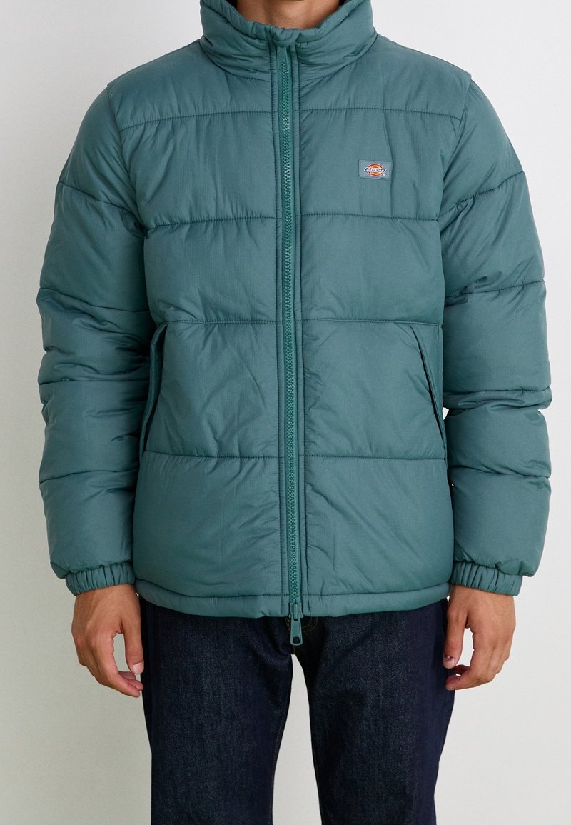 Homme portant une veste matelassée turquoise avec fermeture éclair frontale, poignets élastiques, poches latérales et petit patch logo sur la poitrine.