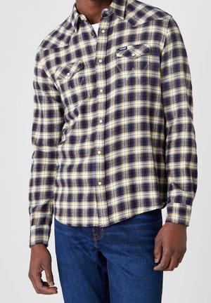 Homme portant une chemise à carreaux jaune, bleue et blanche à boutons avec poches poitrine et un jean bleu foncé, debout devant un fond uni.