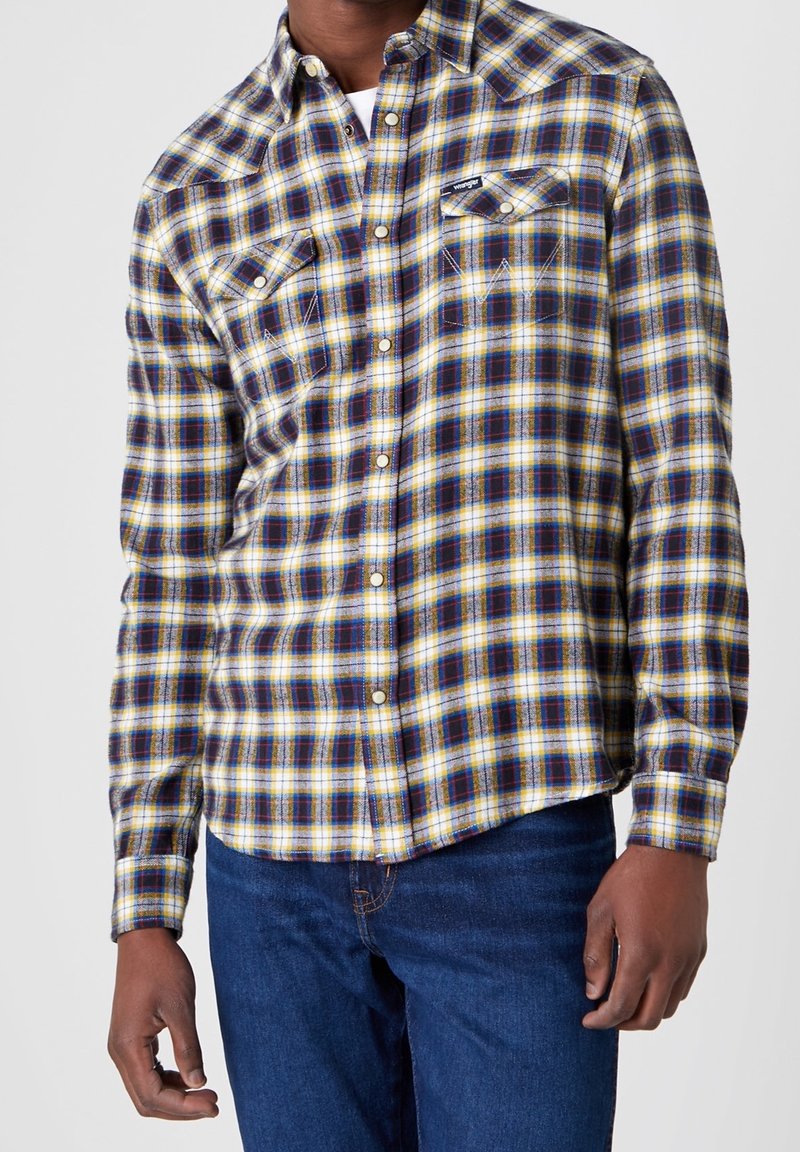 Homme portant une chemise à carreaux jaune, bleue et blanche à boutons avec poches poitrine et un jean bleu foncé, debout devant un fond uni.