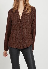 Långärmad brun blus med ett subtilt prickigt mönster, två bröstfickor och en v-ringning, tillverkad av ett mjukt tyg. Kombinerad med svarta byxor.