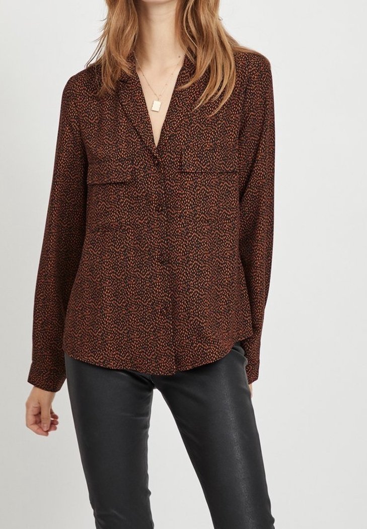 Långärmad brun blus med ett subtilt prickigt mönster, två bröstfickor och en v-ringning, tillverkad av ett mjukt tyg. Kombinerad med svarta byxor.