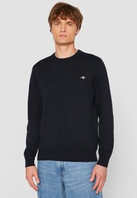 Sötét tengerészkék kötött pulóver kerek nyakkivágással, hosszú ujjakkal és egy kis logóval a mellkasán. Világoskék farmerrel párosítva.