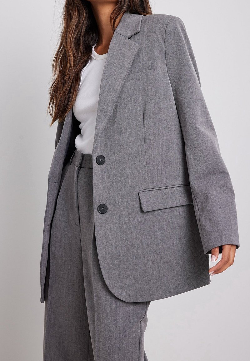 Veste grise sur mesure avec épaulettes structurées, deux boutons noirs et une unique poche poitrine. Portée sur une chemise blanche, avec un pantalon gris assorti.