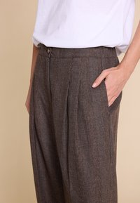 Brune rutete bukser laget av teksturert stoff, med folder og en sidelomme. Knapplukking i midjen.