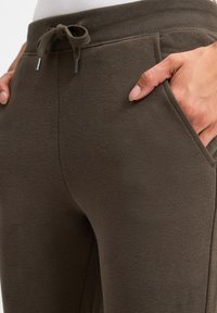 Tmavě olivově zelené tepláky vyrobené z měkkého materiálu s žebrovaným pasem a šňůrkou na stažení. Obsahují boční kapsy a hladkou texturu.