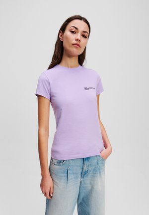 T-shirt lilla in cotone con collo tondo e maniche corte. Presenta un logo nero sul lato sinistro del petto. Abbinato a jeans azzurri chiari.