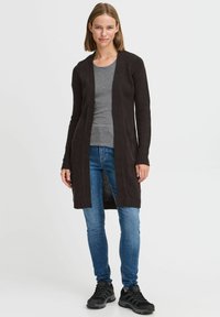 Long, open-front cardigan in dark brown knit material, featuring ribbed cuffs and two front pockets, paired with a gray top and blue jeans.