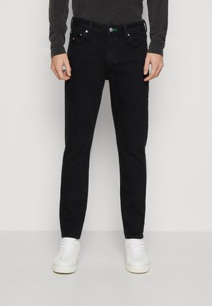Homme portant un jean slim noir et des baskets blanches, debout devant un fond clair uni.