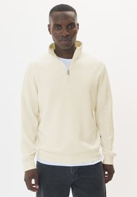Crème-kleurige pullover met een hoge kraag, ritssluiting en lange mouwen. Zachte stof met een gladde textuur, gedragen over een wit T-shirt.