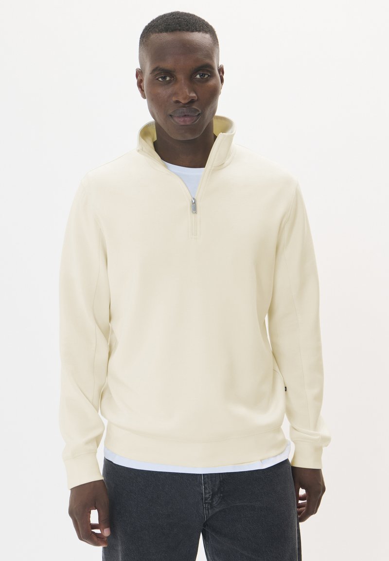 Crème-kleurige pullover met een hoge kraag, ritssluiting en lange mouwen. Zachte stof met een gladde textuur, gedragen over een wit T-shirt.