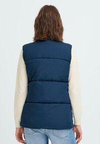 Jūras zilā dūnu veste ar kvadrātveida šuvēm. Gluda tekstūra, bez piedurknēm, pārklāta ar gaišas krāsas garām piedurknēm.