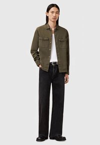 Giovane con capelli neri lunghi, indossante una giacca verde oliva con bottoni sopra una camicia bianca, jeans neri a gamba larga e scarpe nere, in piedi.