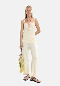Kremowy, dzianinowy top na ramiączkach oraz pasujące szerokie spodnie, do tego naszyjnik z jasnym wiszącym elementem i paskowana torba w kolorze żółto-zielonym.
