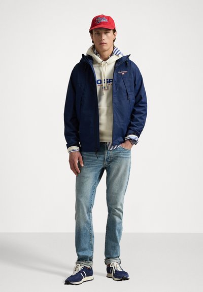 Marineblå vandtæt jakke med hætte, over en beige hættetrøje og blå jeans. Kompletteret med en rød kasket og blå sneakers.