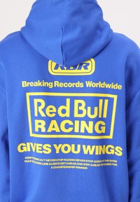 Sudadera azul con un gran gráfico amarillo que dice "Red Bull Racing Te Da Alas" en la parte posterior, junto con texto adicional en un tamaño más pequeño.