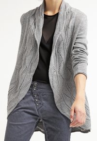 Ljusgrå stickad cardigan med vågmönster, öppen front, långa ärmar och en texturerad yta. Matchad med mörka byxor med knappar.