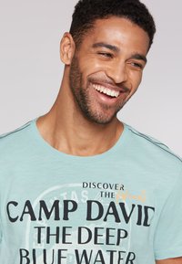 Světle modré bavlněné tričko s černým a oranžovým potiskem. Má kulatý výstřih a krátké rukávy, s jemným černým šitím na ramenech.