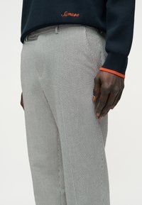 Μαύρο πουλόβερ με πορτοκαλί μπορντούρα και κέντημα "Sansoe", συνδυασμένο με γκρι παντελόνι με κάθετες ρίγες, σε όρθιο άτομο.