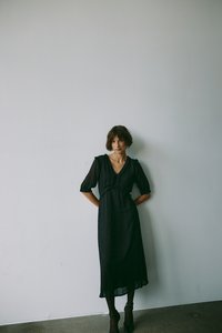 Crna haljina s točkastim uzorkom, kratkim puf rukavima, V-izrezom, stegnutim strukom i midi dužinom, u kombinaciji s crnim potpeticama ispred neutralne pozadine.