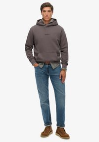 Sudadera gris con bolsillo frontal tipo canguro, sobrecamiseta a cuadros, combinada con jeans de denim azul y botas marrones con cordones.