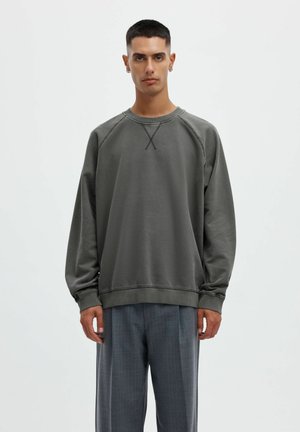 Homme aux cheveux courts portant un sweat-shirt ample gris foncé et un pantalon gris à fines rayures, debout devant un fond clair uni.