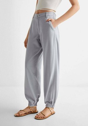 Kobieta ubrana w jasnoszare spodnie dresowe z mankietami, biały krótki top na ramiączkach i złote płaskie sandały, stojąca na tle jednolitego tła.