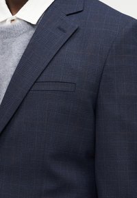 Κοντινό πλάνο ενός μπλε σκούρου καρό σακακιού που φοριέται πάνω από λευκό πουκάμισο με γιακά και γκρι πλεκτό πουλόβερ.