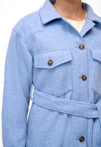 Modrý kabát z vlněné směsi s dvěma kapsami na hrudi, knoflíkovým zapínáním a ladicím páskem. Materiál má jemný, strukturovaný povrch.