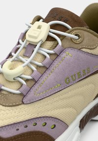 Ανοιχτό καφέ αθλητικό παπούτσι με μοβ και καφέ λεπτομέρειες. Διαθέτει υφασμάτινο επάνω μέρος, υφές σε πάνελ, ελαστικά κορδόνια και λαστιχένια σόλα. Ορατό λογότυπο.