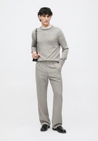 Suéter cinza claro com decote redondo, combinado com calças pregueadas cinza claro e ajustadas. O modelo usa sapatos pretos e segura uma mala de ombro.