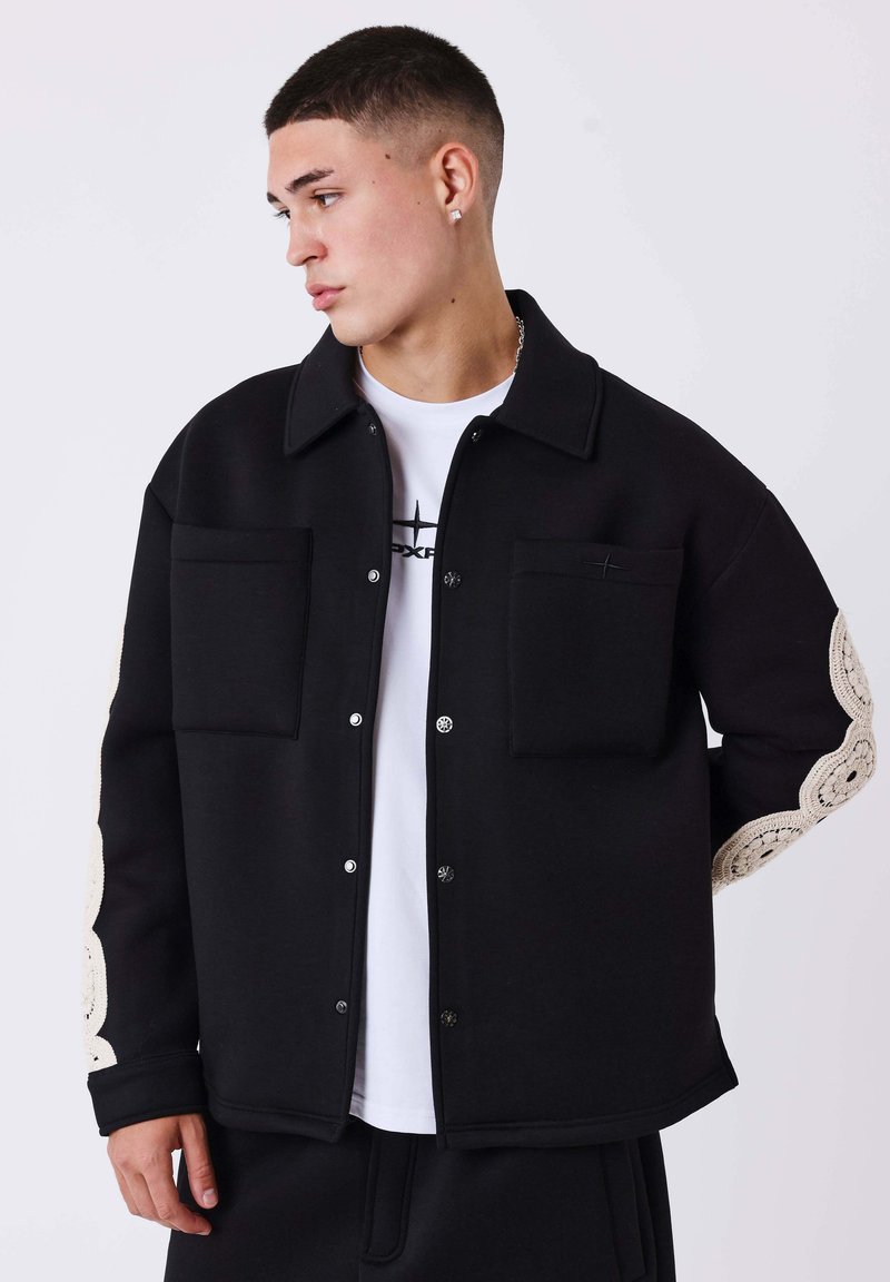 Veste noire avec deux poches poitrine, boutons-pression, et un accent unique à motif crème sur les manches. Portée par-dessus une chemise blanche.