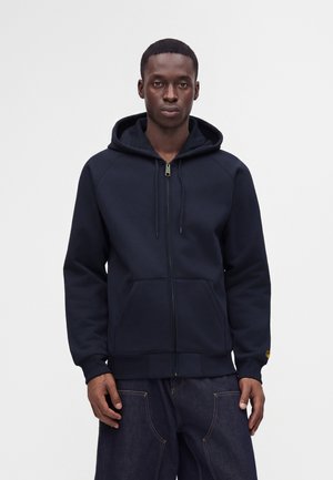 Homme portant un sweat à capuche bleu marine zippé avec poches avant et un jean bleu foncé, debout devant un fond clair uni.