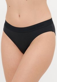 Zwarte bikini briefs met een gladde textuur en een elastische tailleband met het reliëf "Organic Basics"-merk. Minimalistisch ontwerp, middelhoog uitgesneden.