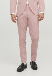 Svijetlo ružičaste hlače krojene u slim fit stilu, izrađene od glatkog materijala, sa bočnim džepovima i suptilnim plitkim naborima, uparene s crnim loafers cipelama.