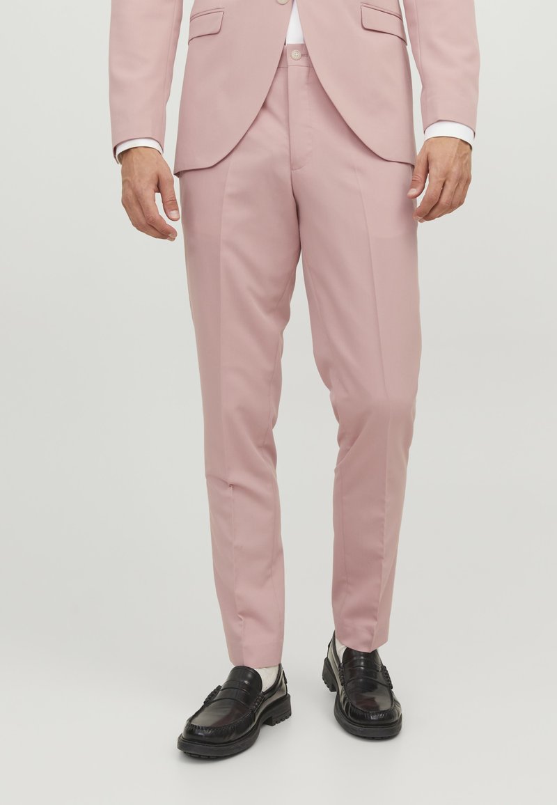 Svijetlo ružičaste hlače krojene u slim fit stilu, izrađene od glatkog materijala, sa bočnim džepovima i suptilnim plitkim naborima, uparene s crnim loafers cipelama.