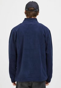 Pullover in pile di colore blu navy con colletto alto, maniche lunghe e vestibilità rilassata. Texture liscia senza hardware visibili o motivi.