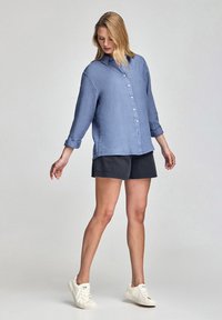 Ljusblå skjorta med knappar och långa ärmar, ihopparad med mörka shorts och vita sneakers. Skjortan har en strukturerad tygkvalitet och en klassisk krage.