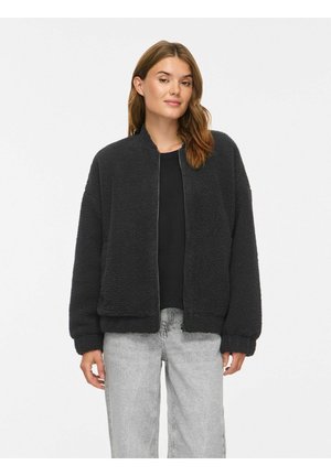 Kvinde iført en sort fleecejakke med lynlås, sort skjorte og lysegrå jeans, stående foran en ensfarvet hvid baggrund.