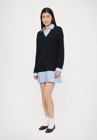 Fekete kötött pulóver V-nyakkivágással, világoskék csíkos ingruha fölött. A modell fehér zoknit és sötét bordó cipőt visel.