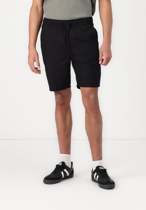 Shorts en coton noir avec taille élastique, poches latérales et coupe décontractée, associés à des baskets noires et des chaussettes blanches montant à la cheville.