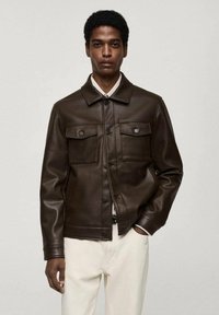 Veste en cuir marron avec un col, deux poches poitrine et des fermetures à boutons, assortie à un pantalon de couleur claire. Texture lisse.