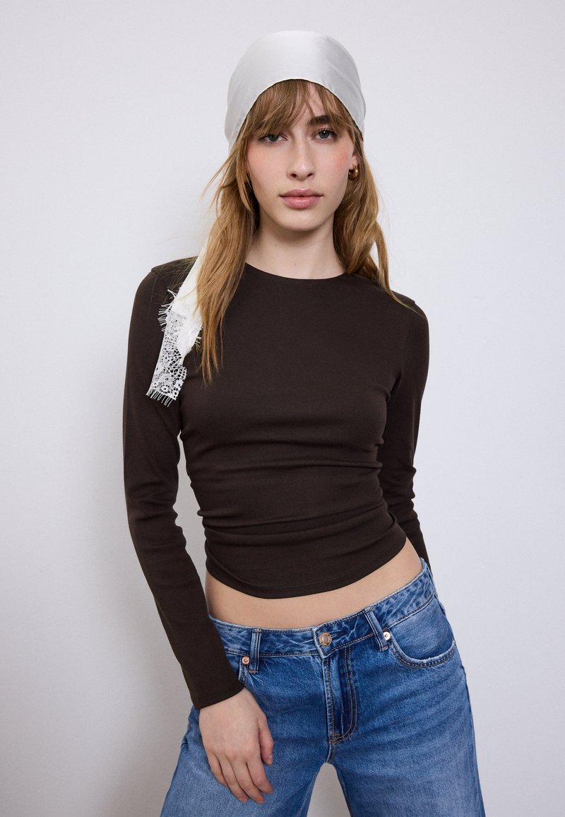 Donna con capelli castano chiaro che indossa un foulard bianco, una maglia nera aderente a manica lunga e jeans blu su uno sfondo chiaro semplice.